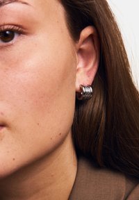 Orecchino a cerchio in argento con una fascia texturizzata e piccoli embellimenti, indossato all'orecchio del modello su uno sfondo neutro.