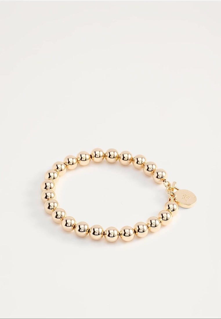 Bracelet extensible en perles dorées avec des perles rondes et un petit pendentif rond gravé d'initiales sur fond blanc.