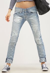 Jeans de mezclilla azul claro desgastados con acentos deshilachados y rotos. Corte ajustado, con dobles en los tobillos, combinados con zapatillas negras.