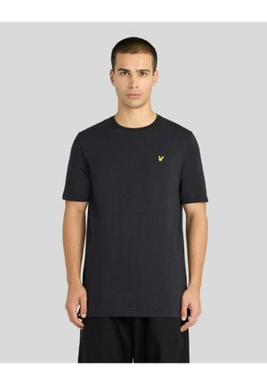 Ung mand vendt fremad iført en ensfarvet sort kortærmet T-shirt med en lille gul fuglelogo på brystet mod en grå baggrund.