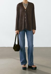 Person wearing brown buttoned cardigan, blue jeans, platform sandals, holding a black handbag against a plain background.