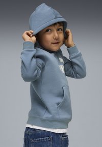 Sweatshirt à capuche bleu clair avec une poche frontale, arborant un logo blanc. Porté par-dessus une chemise blanche et associé à un jean bleu foncé.