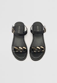Schwarze flache Sandalen mit goldener Kettenverzierung. Mit einem Knöchelriemen und einer strukturierten Sohle. Das Design ist minimalistisch und modern.