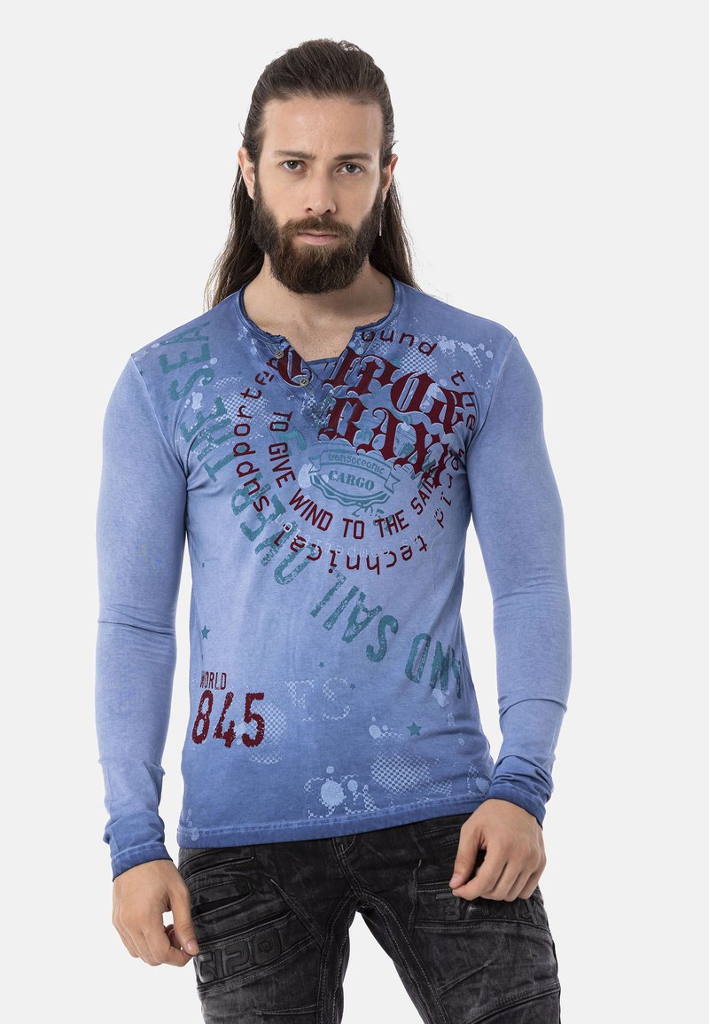Blaues Langarmhemd mit Rundhalsausschnitt, das verschiedene grafische Drucke in Rot und Grün zeigt. Der Stoff hat eine weiche Textur mit verblassten Bereichen.