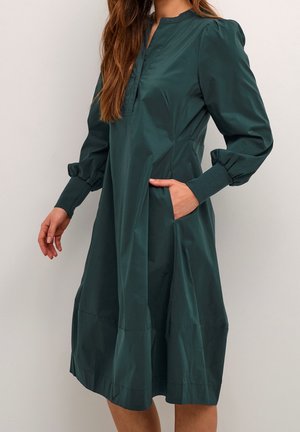 Frau trägt ein langärmeliges, dunkelgrünes, knielanges Kleid mit gerafften Bündchen und einer Hand in einer seitlichen Tasche.