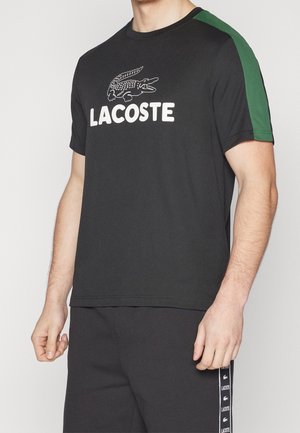 Mann trägt schwarzes Lacoste-T-Shirt mit grünen Schulterstreifen und schwarze Hose mit Lacoste-Logo-Band an der Seite.