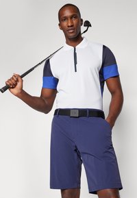 Bílá a námořnická polo tričko s modrými akcenty, se zipovým uzávěrem, spárované s námořnickými šortkami a černým opaskem s logem. Golfová hůl držena v rukou.
