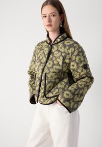 Veste matelassée de couleur vert olive avec un motif géométrique, dotée d'un col montant, d'une bordure noire et de poches avant. Matériau léger, design décontracté.