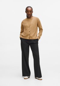 Beige kardigán V-nyakkal és gombos záródással, magas derekú fekete nadrággal párosítva. A modell kezeit a zsebében tartja, fehér cipőt visel.