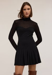 Černé šaty s přiléhavým, texturovaným topem s dlouhým rukávem a skládací sukní. Vyrobeno z pleteniny, která se vyznačuje hladkým povrchem.