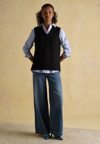 Junge Frau steht mit den Händen hinter dem Rücken, trägt einen schwarzen ärmellosen Pullover über einem gestreiften Hemd, weite Jeans und spitze Schuhe.