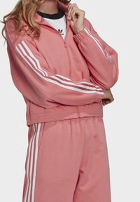 Giacca sportiva rosa a maniche lunghe, con strisce bianche sulle spalle e chiusura con zip. Tessuto morbido, orlo a coste, abbinata a pantaloni coordinati.