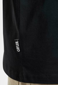 Sort bomulds t-shirt med lige kant. Har et hvidt stofmærke med "CAYLER" syet på venstre side. Glat tekstur, minimalistisk design.