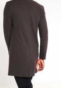 Brun uldfrakke med en skræddersyet pasform, enkelt ventil og knapmanchetter, der viser en glat tekstur og minimalistisk design fra ryggen.