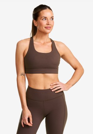 Kvinna i brun sport-bh och leggings poserar med ena handen på höften, ser svagt åt sidan och visar en sol-tatuering på vänster arm.