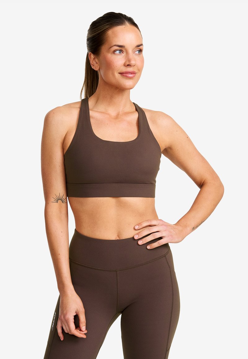 Kvinna i brun sport-bh och leggings poserar med ena handen på höften, ser svagt åt sidan och visar en sol-tatuering på vänster arm.