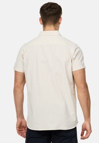 Camisa de manga corta en tela beige claro, con cuello y líneas limpias. La parte trasera tiene una costura en el yugo y es suave con un minimalismo en los detalles.