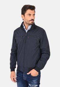 Giacca leggera di colore blu navy con tasche con zip, colletto alto, polsini a costine e orlo; texture morbida, design casual.