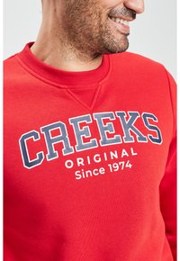 Sudadera roja con cuello redondo, que presenta "CRAEKS" en letras negritas blancas y azules, y "ORIGINAL Desde 1974" impreso debajo. Textura suave.