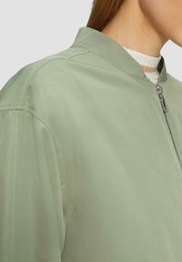 Giubbotto bomber verde chiaro con colletto alto, chiusura con zip e tasche laterali. Il tessuto appare liscio con dettagli di cucitura visibili.
