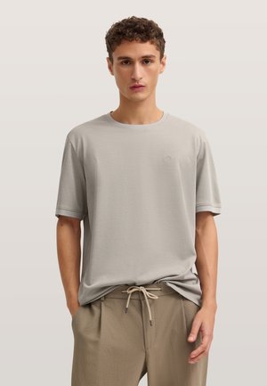 Junger Mann mit kurzen lockigen Haaren, der ein hellgraues T-Shirt mit Rundhalsausschnitt und beige Kordelzughosen trägt, die Hände in den Taschen.