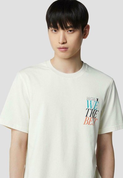 Jeune homme aux cheveux noirs portant un t-shirt blanc avec "MCM x WE THE BEST" imprimé sur la poitrine gauche.