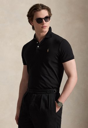 Jeune homme portant un polo noir, un pantalon noir, des lunettes de soleil et une montre-bracelet, une main dans la poche, regardant sur le côté.