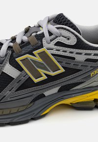 Gros plan d'une chaussure de course noire, grise et jaune avec des lacets gris et un grand logo stylisé "N" sur le côté.