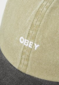 Grønn og svart caps laget av bomull, med en glatt tekstur. "OBEY" brodert i hvitt på frontpanelet, med buet skygge.