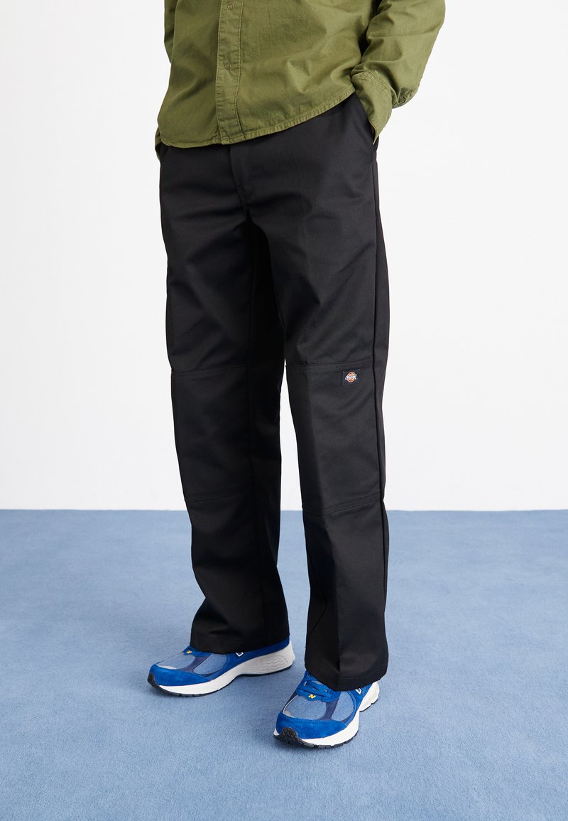 Black pants with a smooth texture, featuring knee panels and a small logo patch. Paired with blue athletic shoes on a light blue surface.