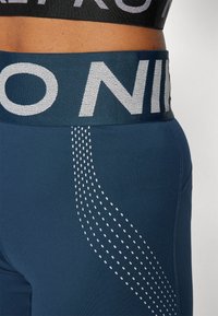Mėlynos sportinės leginsai su plačia elastinga juosta, ant kurios parašyta "NIKE" pilkšva spalva. Šoninio rašto dekoras sudarytas iš mažų baltų taškelių, sudarančių bangų dizainą.