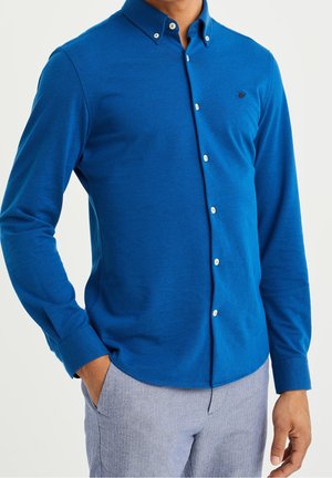 Mies, jolla on istuva sininen nappipaita valkoisilla napeilla ja vaaleanharmaat housut, seisoo yhdellä kädellä taskussa.