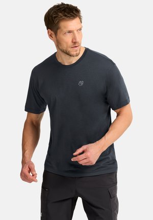 Man wearing dark blue short-sleeve shirt and black pants looks right with hands slightly raised against plain background.