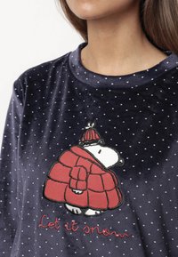 Camiseta de terciopelo azul marino con lunares blancos, que presenta un personaje bordado con un abrigo rojo y el texto "Que nieve" en hilo rojo.