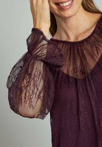 Blusa a maniche lunghe in pizzo viola scuro con maniche a sbuffo trasparenti e scollatura rotonda. Motivo floreale a rilievo su tutto il tessuto.
