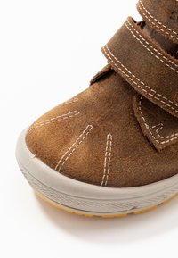 Zapato de ante marrón con punta redonda, suela de goma gris y costuras crema. Cuenta con dos correas de Velcro ajustables para el cierre.