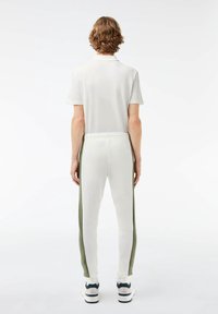 Bílé polo tričko s krátkým rukávem a kalhoty s elastickým pasem, které mají zelené boční pruhy, a bílé tenisky. Hladká tkanina, přiléhavý střih.