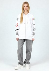 Felpa bianca con cerniera e vari disegni grafici, abbinata a jeans grigi larghi e sneakers bianche. Tessuto liscio, design casual.