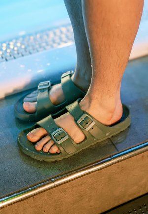 Voeten met natte olijfgroene gespsandalen staan op natte grond vlakbij een metalen rooster oppervlak.