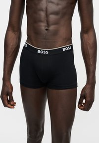Mužský trup oblečený v čiernych boxerkoch s bielym logom "BOSS" na pásiku na bielom pozadí.