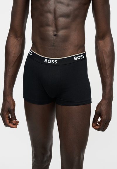 Torse masculin portant un boxer noir avec un logo blanc "BOSS" sur la ceinture, sur fond blanc uni.