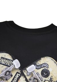 Černé tričko s kulatým výstřihem, které zobrazuje grafiku dvou skateboardových desek, texturovaných koleček a různých samolepek v krémové a černé barvě.