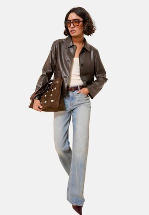Vrouw met een oversized bruine leren jas, lichtblauwe spijkerbroek, beige top, grote bruine handtas, bruine zonnebril en paarse puntige schoenen.