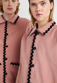 Ružový látkový kabát s čiernym vrúbkovaným lemovaním pozdĺž prednej strany, veľkými čiernymi gombíkmi a vreckom na ľavej strane.