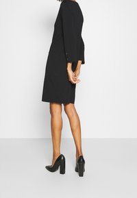 Robe noire à manches trois-quarts, silhouette droite et accents de boutons. Portée avec des chaussures à talons hauts noires, texture lisse.