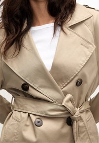 Kobieta ubrana w beżowy, podwójnie zapięty trencz związany w talii na białej, prążkowanej koszuli, z luźnymi, ciemnobrązowymi włosami.