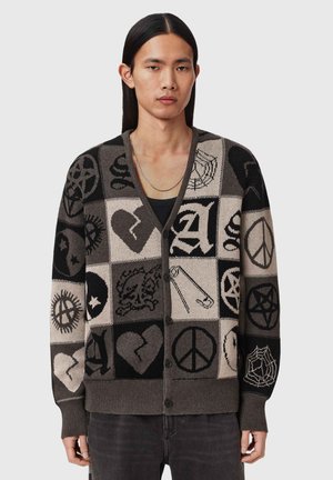 Młody mężczyzna z długimi czarnymi włosami, ubrany w rozpinany kardigan z czarnymi, szarymi i beżowymi motywami patchworkowymi, w tym sercami, znakami pokoju i pająkami.