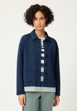 Kobieta z krótkimi kręconymi włosami, ubrana w granatową zapinaną na guziki kurtkę na koszulę w paski i jasnozielone spodnie, stojąca na białym tle.