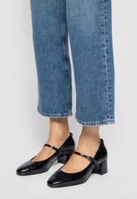 Sorte lakk lærblokksko med spiss tå og tynn ankelrem, med krokodilleprøvd tekstur. Bært med avkappede blå jeans.