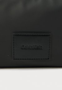 Černá nylonová taška s texturovanou koženou etiketou s vyráženým brandingem "Calvin Klein", minimalistickým designem a elegantním obdélníkovým tvarem.
